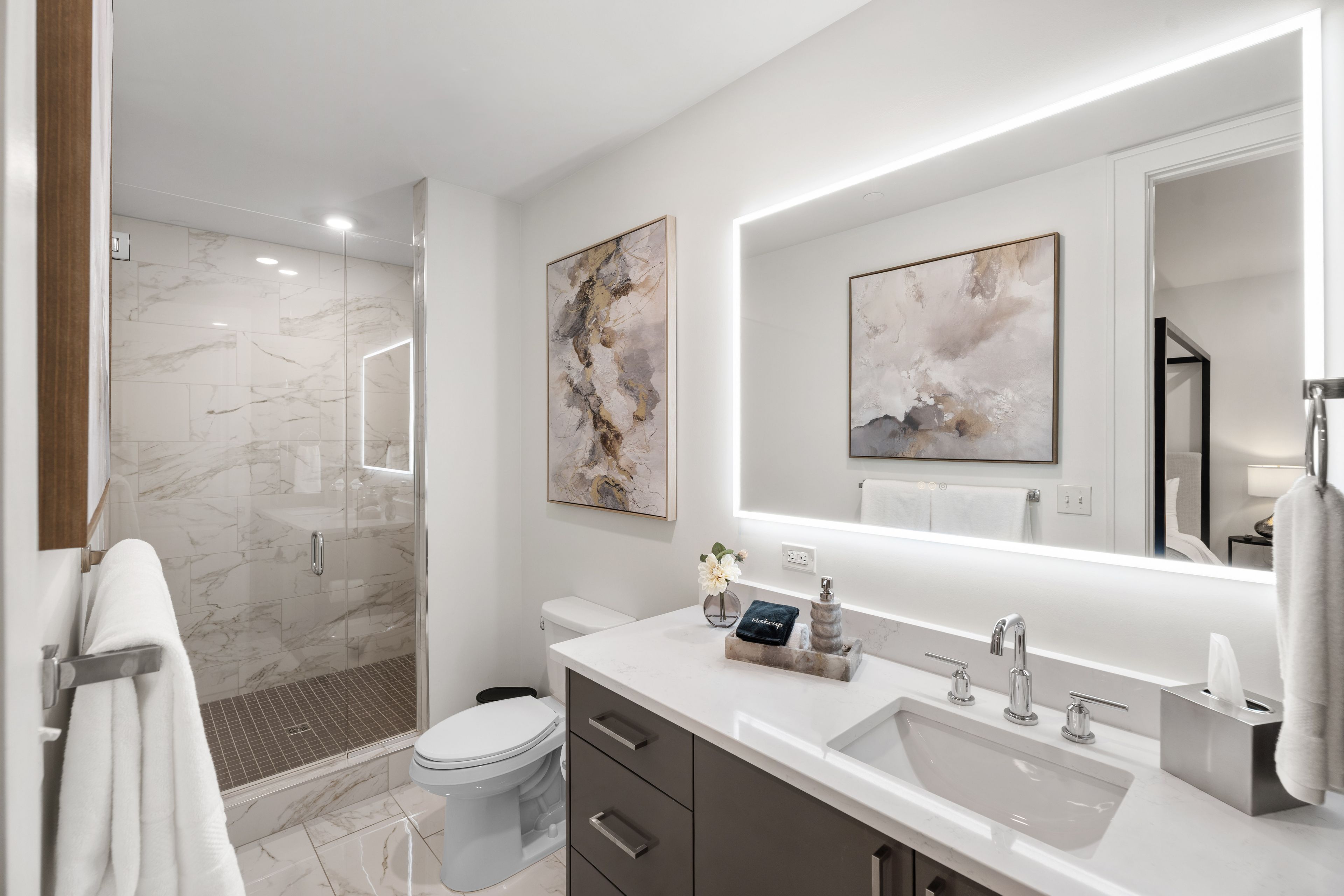 Een moderne badkamer met een douchecabine van glas, een toilet, een wastafel met een marmeren blad en twee abstracte schilderijen aan de wanden.