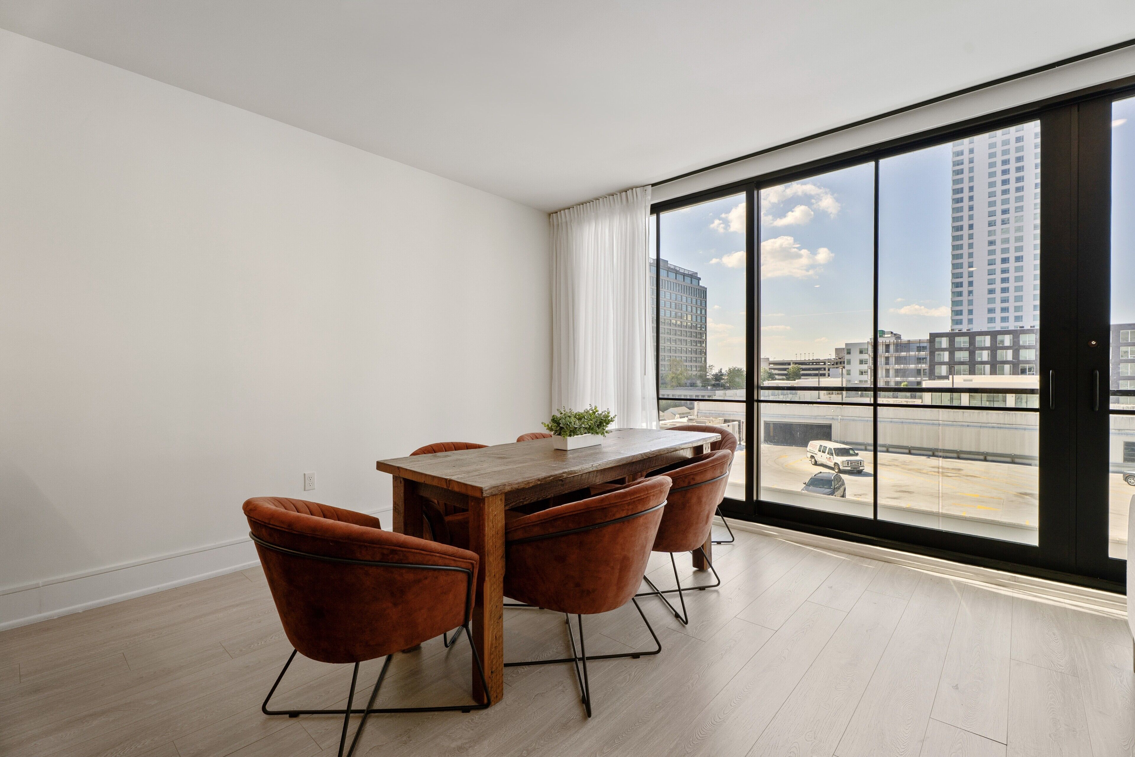 A modern dining area with a wooden table and four chairs, large windows with a city view, and a clean, white interior.