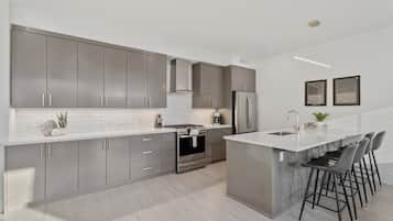 A modern kitchen with grey cabinets, a central island with bar stools, stainless steel appliances, and a white backsplash.