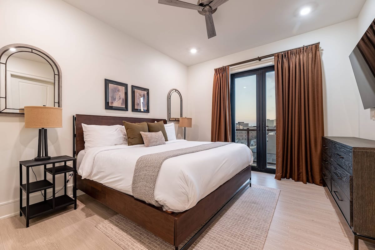 Una habitación de hotel moderna con una cama grande, una mesita de noche con lámpara, un aparador y un amplio ventanal con cortinas.