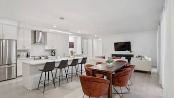 A modern kitchen with white cabinetry, a central island with bar stools, a dining table with chairs, and a flat-screen TV mounted on the wall.