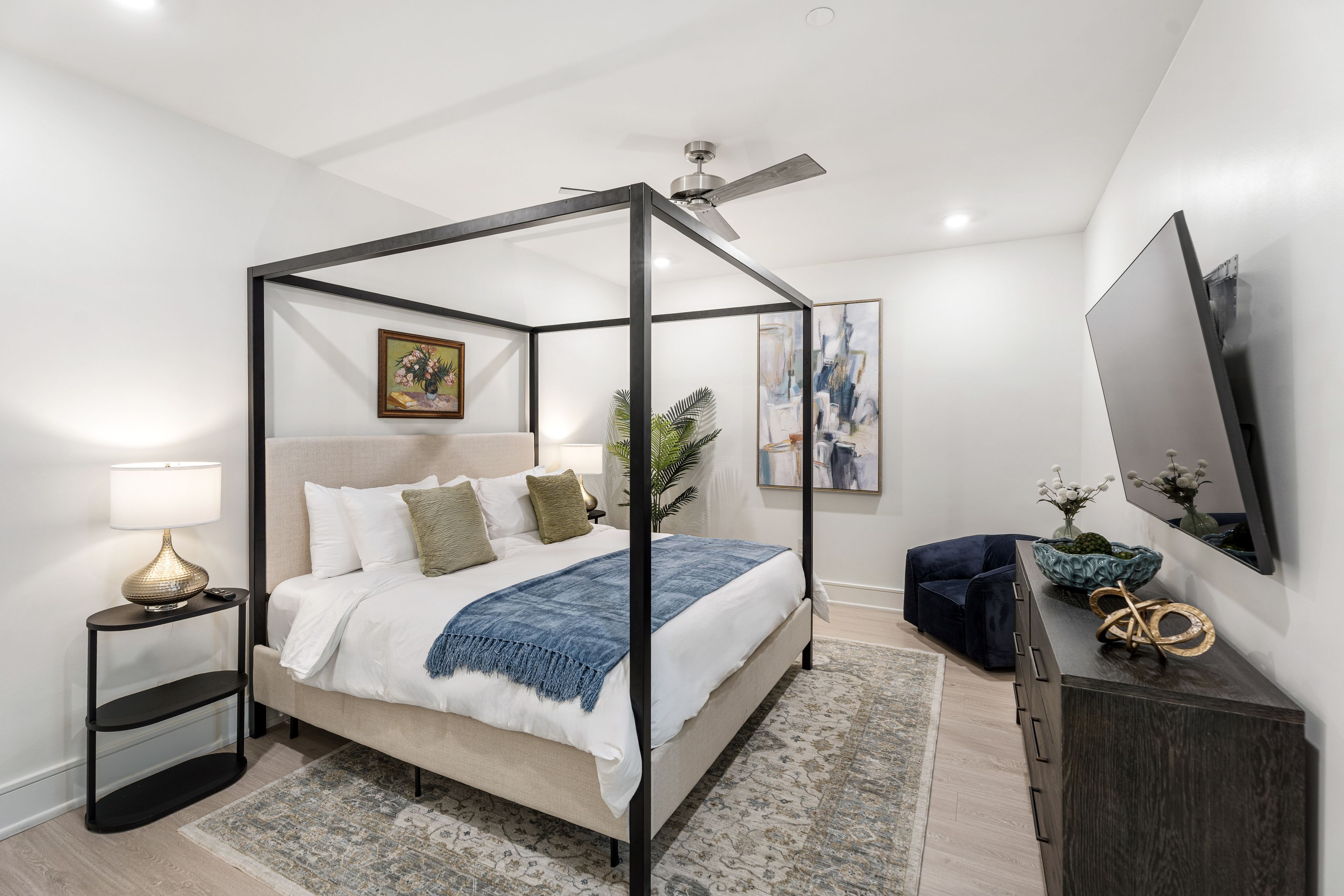 A modern bedroom with a four-poster bed, a ceiling fan, a bedside table with a lamp, a dark blue chair, a dresser, and a large mirror.