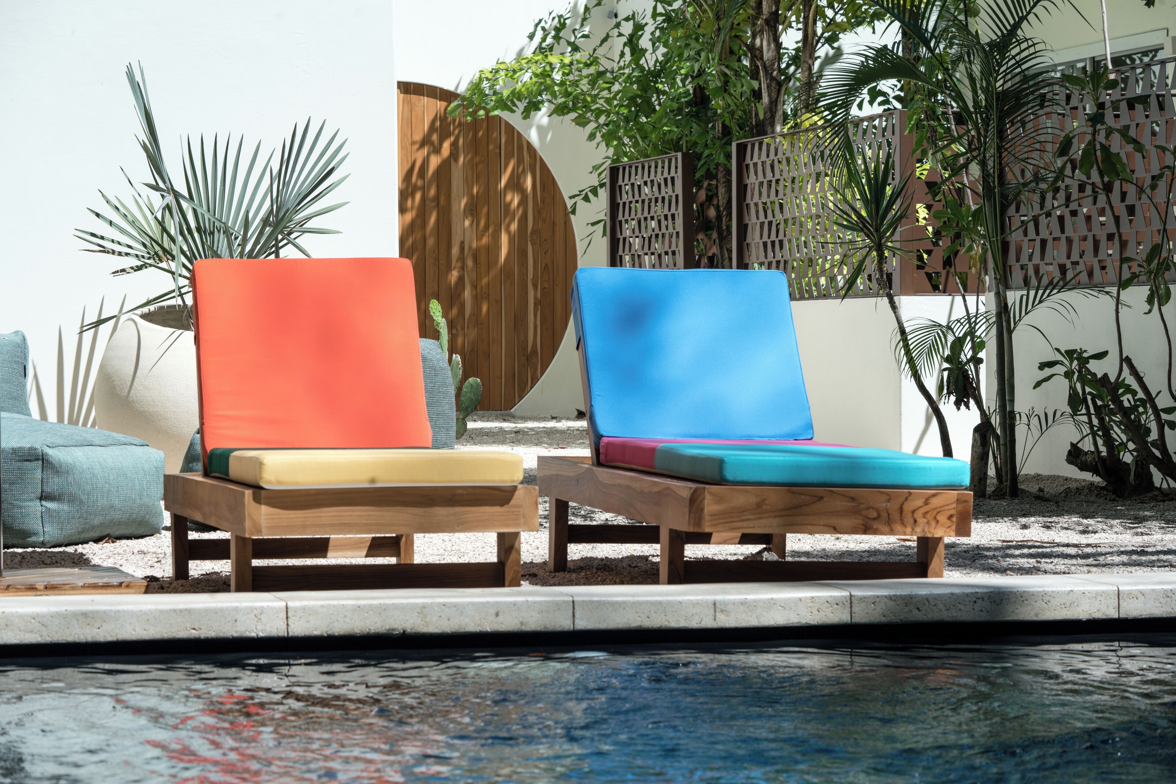 Two wooden lounge chairs with cushions by a poolside, surrounded by plants and a wooden fence.