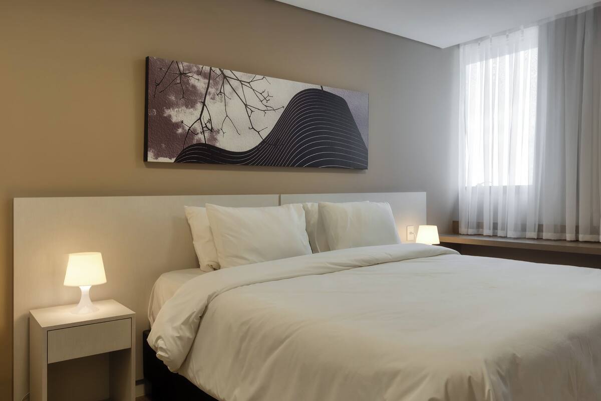 A hotel room with a large bed, two bedside lamps, a nightstand, and a window with sheer curtains.