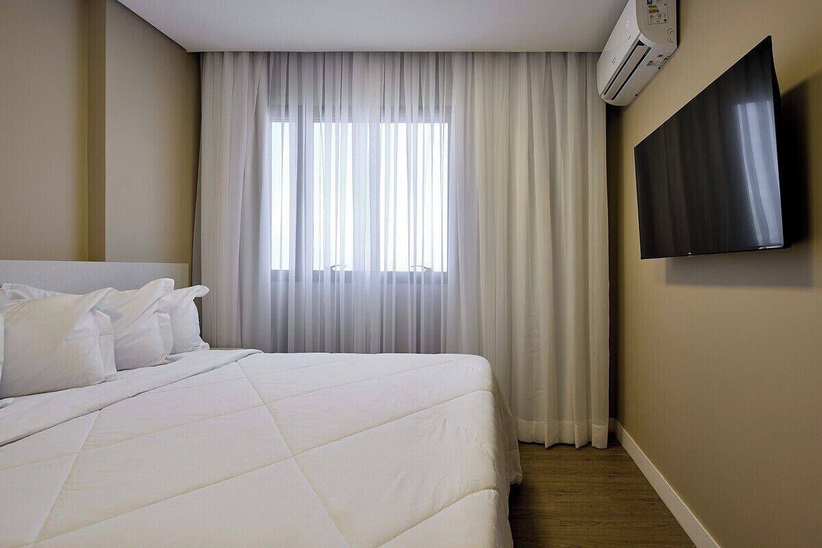 A hotel room with a bed, white curtains, a mounted TV, and an air conditioning unit.