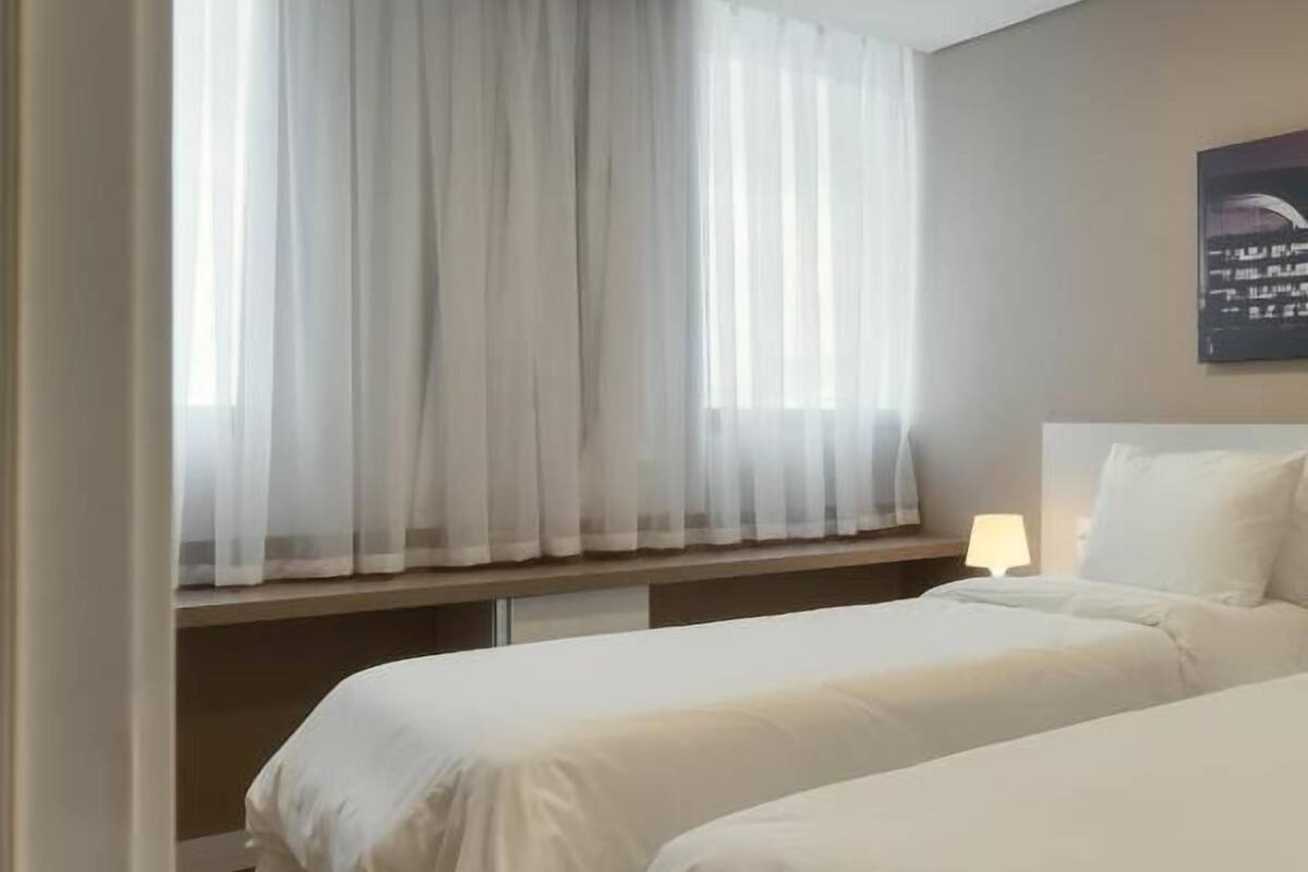 A hotel room with two beds, a window with sheer curtains, a bedside table with a lamp, and a wall-mounted artwork.