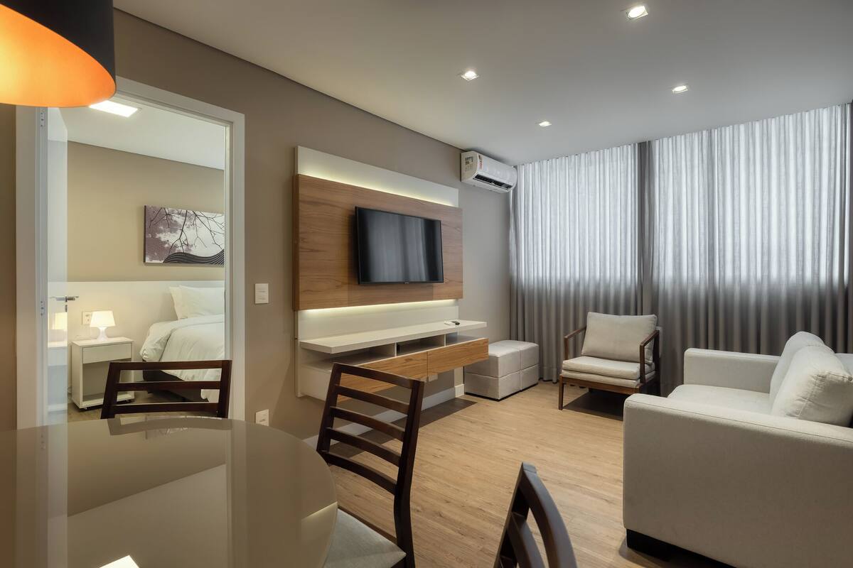 Quarto de hotel moderno com televisão de tela plana presa a um painel de madeira, uma mesa de jantar com cadeiras, um sofá e uma janela grande com cortinas.