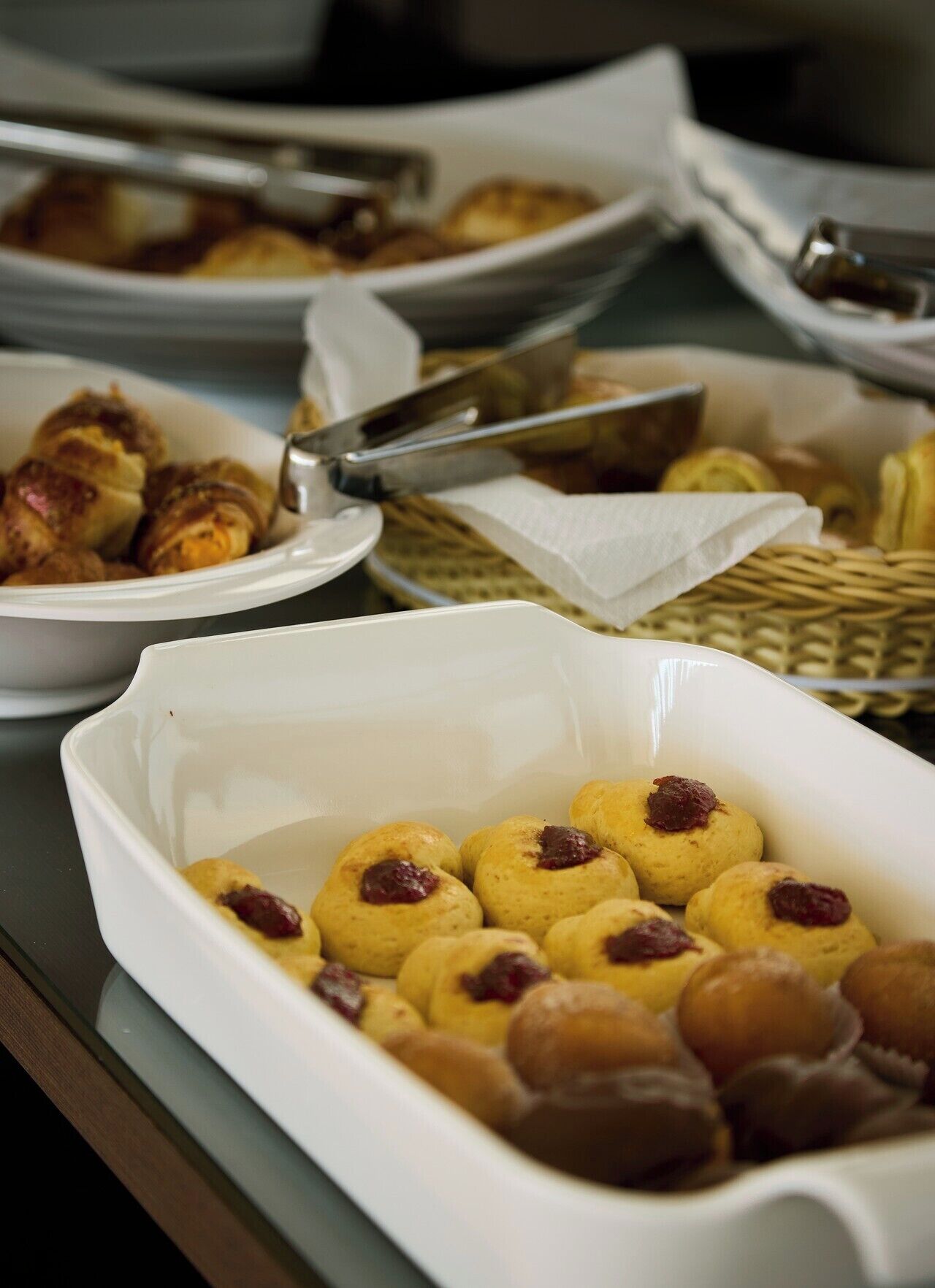 Uma mesa de buffet com diversos alimentos, incluindo pastelaria e um prato branco com pastelaria redonda coberta com um produto vermelho.