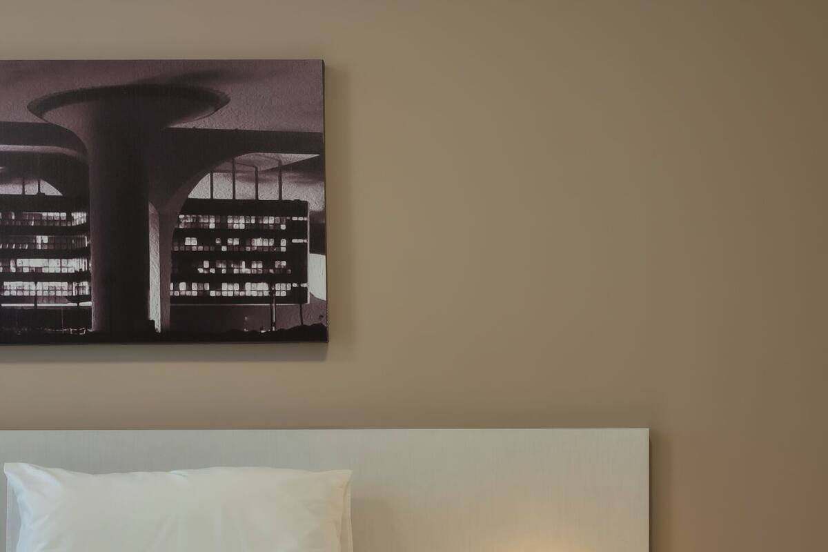 A hotel room with a bed, a nightstand with a lamp, and a cityscape painting on the wall.