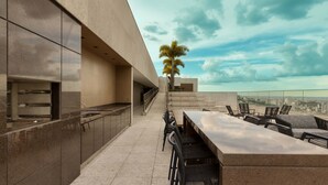 An outdoor kitchen area with a long countertop and bar stools, a palm tree, and a city view. - Palazzo Torquetti Condo Hotel e Eventos (Belo Horizonte)