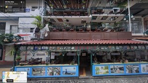 Un bâtiment à deux étages, doté d’un toit recouvert de tuiles, de balcons ornés de plantes et d’un magasin au rez-de-chaussée avec un comptoir bleu où sont exposées des images.
