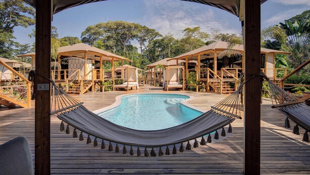 A wooden deck with a pool, hammocks, and wooden structures with thatched roofs.