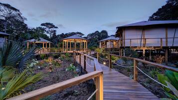 Une terrasse en bois mène à plusieurs constructions également en bois, dotées de murs en verre, et l’ensemble est entouré de plantes tropicales.