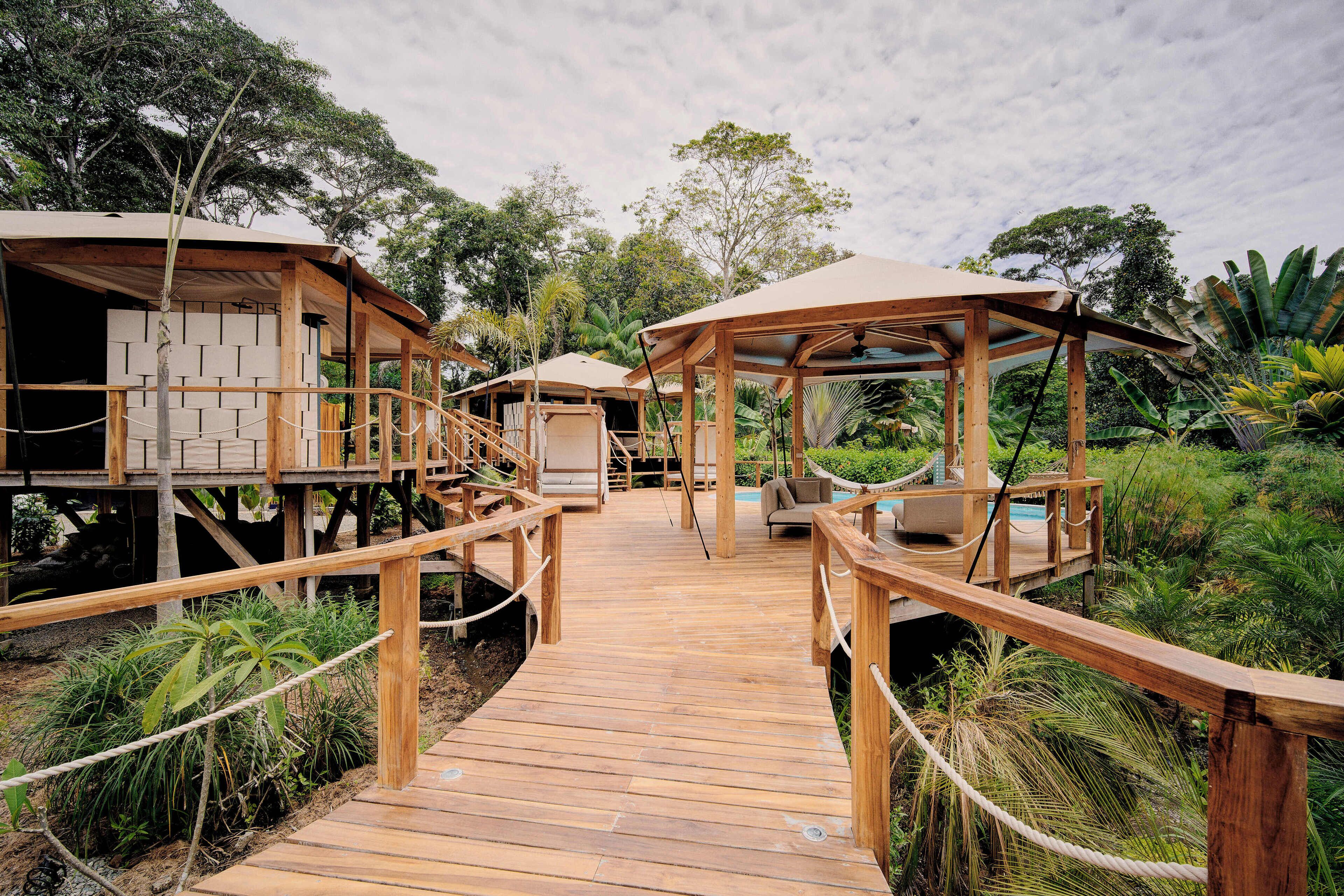 A wooden deck connecting two wooden structures with outdoor seating and a pool area, surrounded by lush greenery.