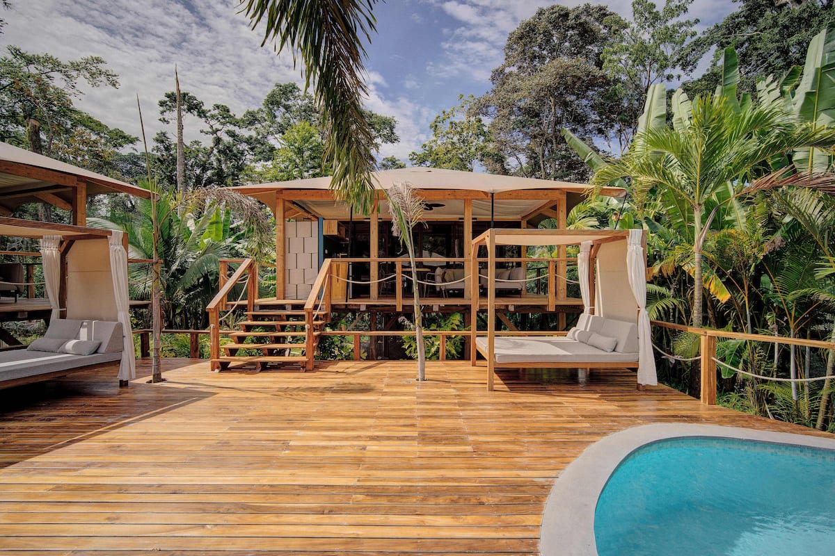 Une terrasse en bois avec une piscine, un espace salon couvert et une végétation luxuriante.