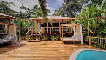 A wooden deck with a pool, a covered seating area, and lush greenery.