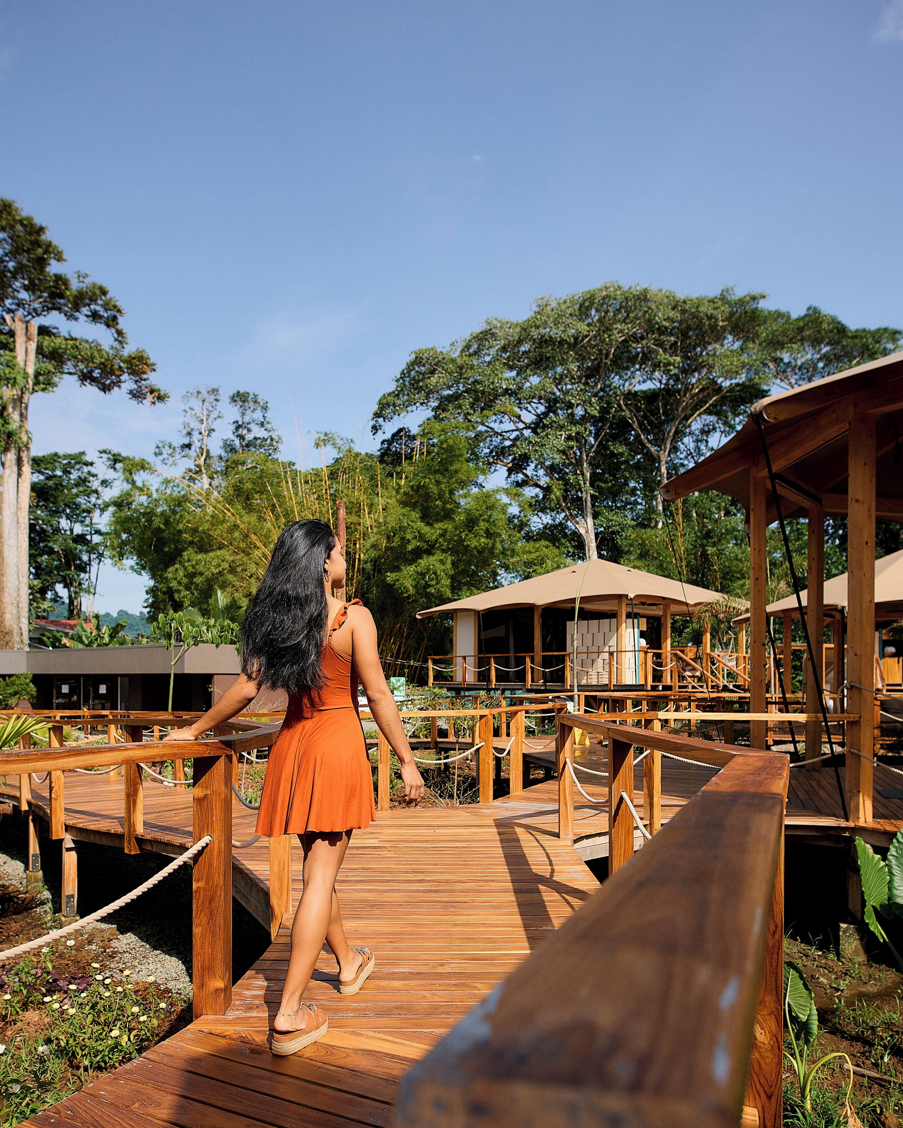 A wooden deck with a woman in an orange dress walking towards a wooden structure with a roof.