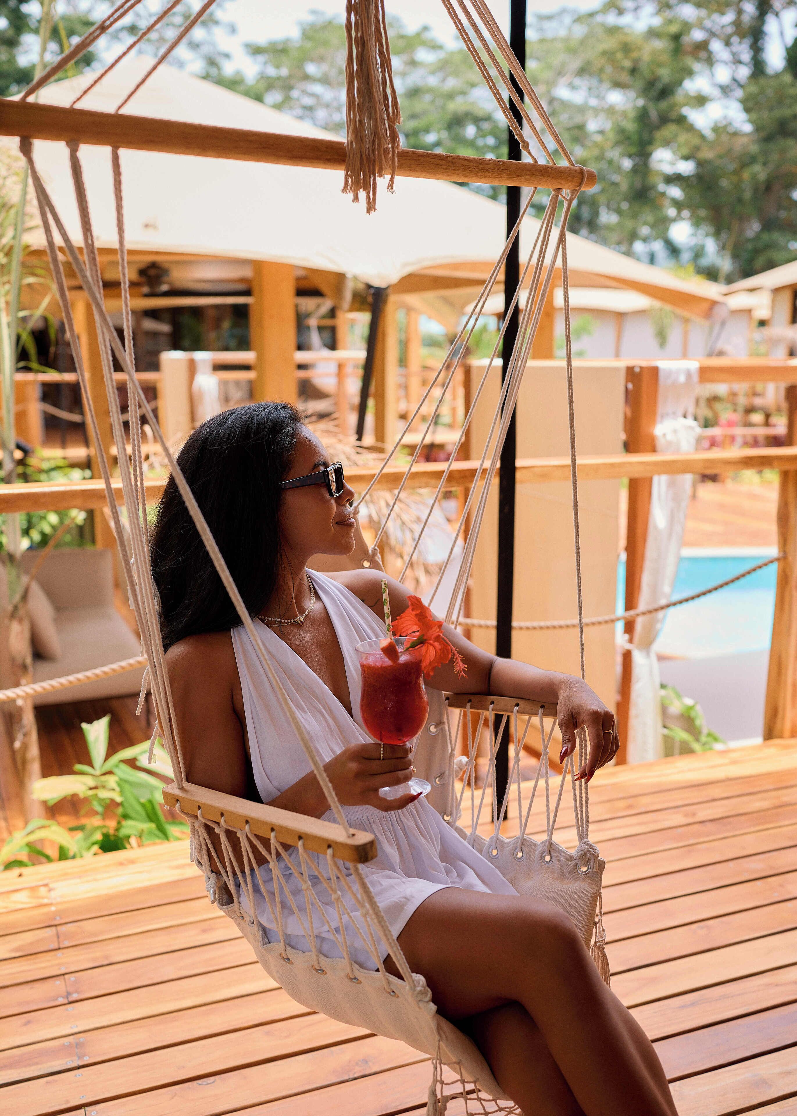 A person relaxing in a hammock chair with a drink, overlooking a pool area.
