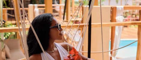 A person relaxing in a hammock chair with a drink, overlooking a pool area.