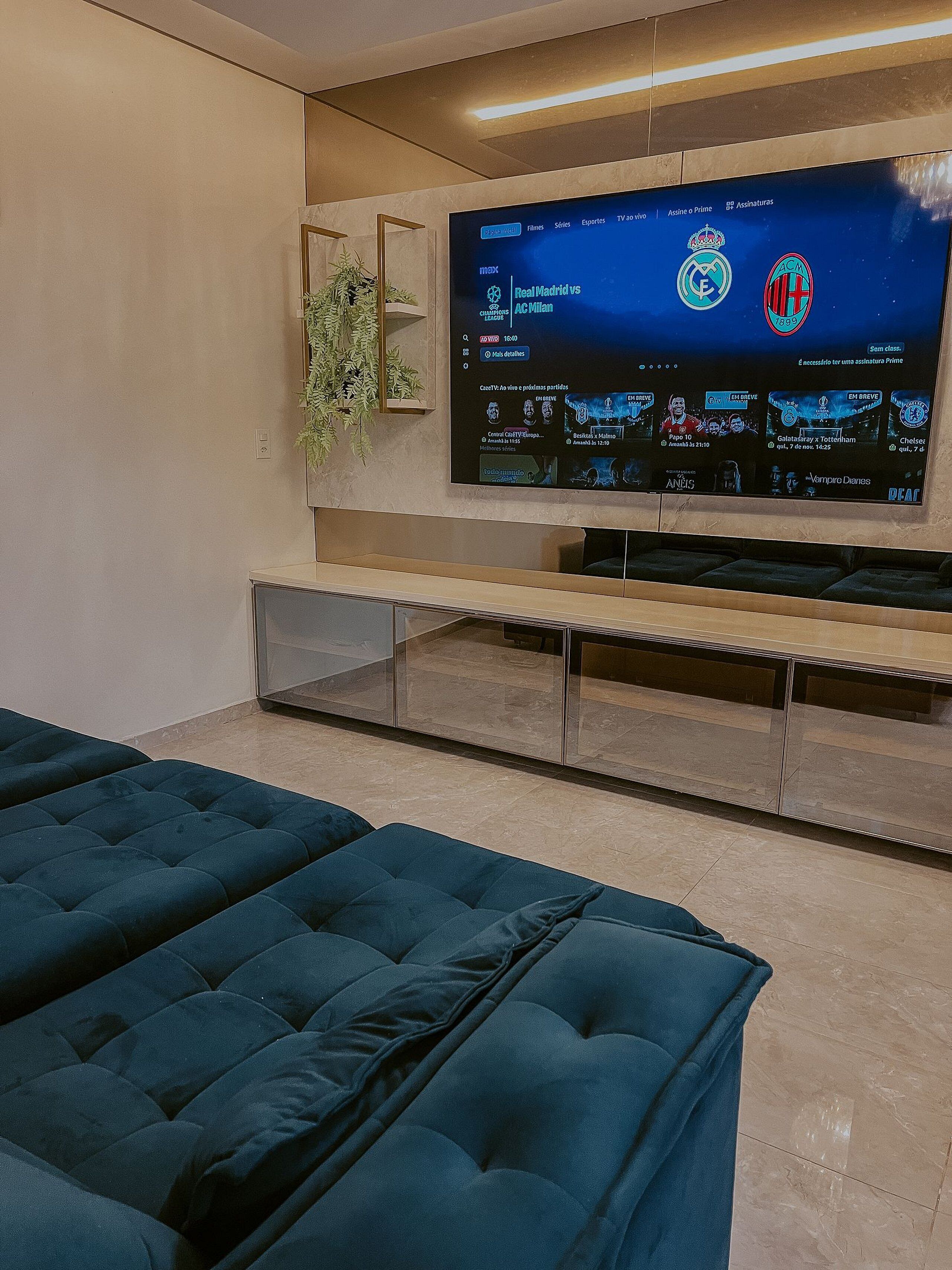A modern living room with a large flat-screen TV mounted on the wall displaying a sports channel. There are two dark blue sofas arranged in the foreground.