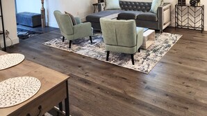 Ein modernes Wohnzimmer mit einem grauen Sofa, zwei Sesseln, einem Holztisch und einem gemusterten Teppich. Ein Beistelltisch ist mit Dekorationen geschmückt, an der Wand hängt ein gerahmtes Kunstwerk und ein Fenster mit Jalousien.