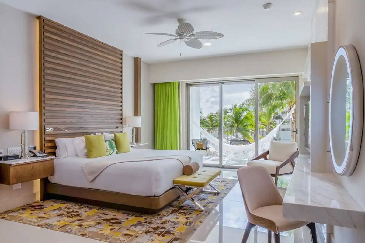 A modern hotel room with a large bed, a desk, a chair, a ceiling fan, and a view of the outdoors.