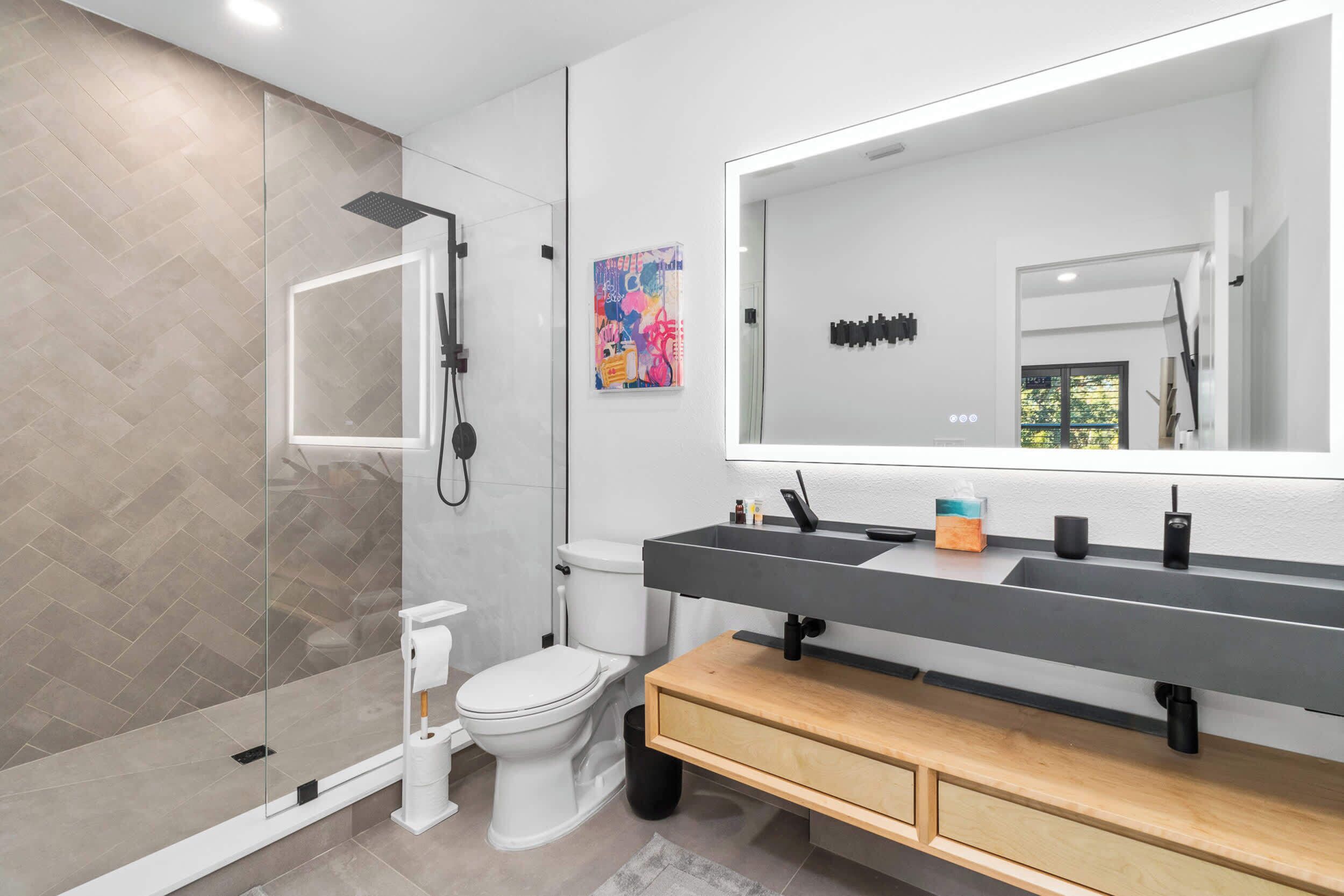A modern bathroom with a glass-enclosed shower, a white toilet, a large mirror, and a wooden vanity.