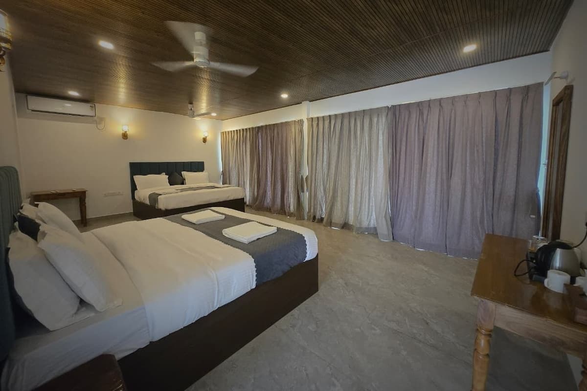 A hotel room with two beds, a wooden ceiling, a ceiling fan, a table with a kettle, and a window with curtains.