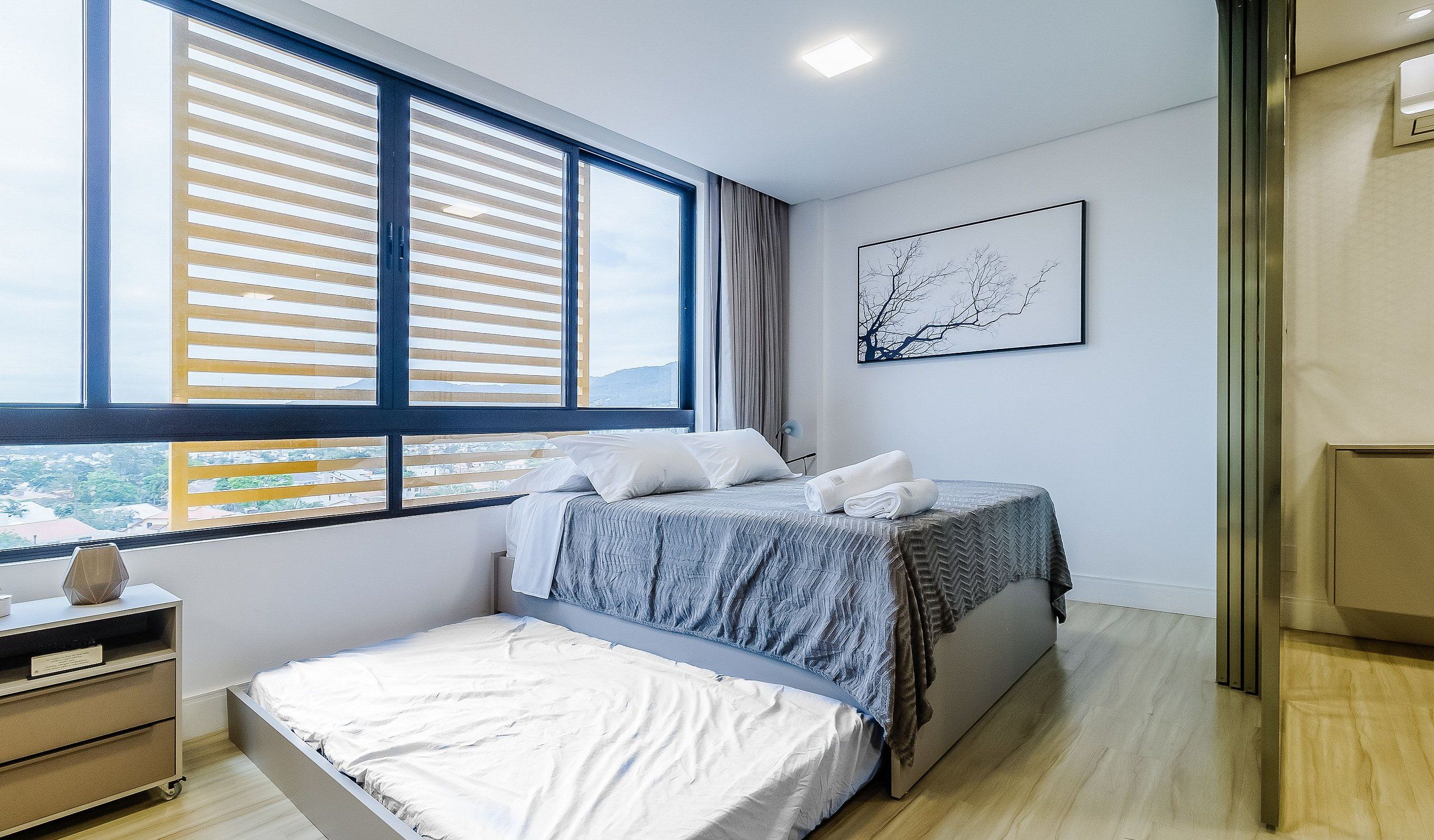 A bedroom with a bed, a nightstand, a window with blinds, a wall-mounted artwork, and a view of the outside.