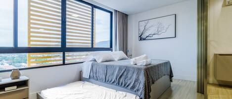 A bedroom with a bed, a nightstand, a window with blinds, a wall-mounted artwork, and a view of the outside.