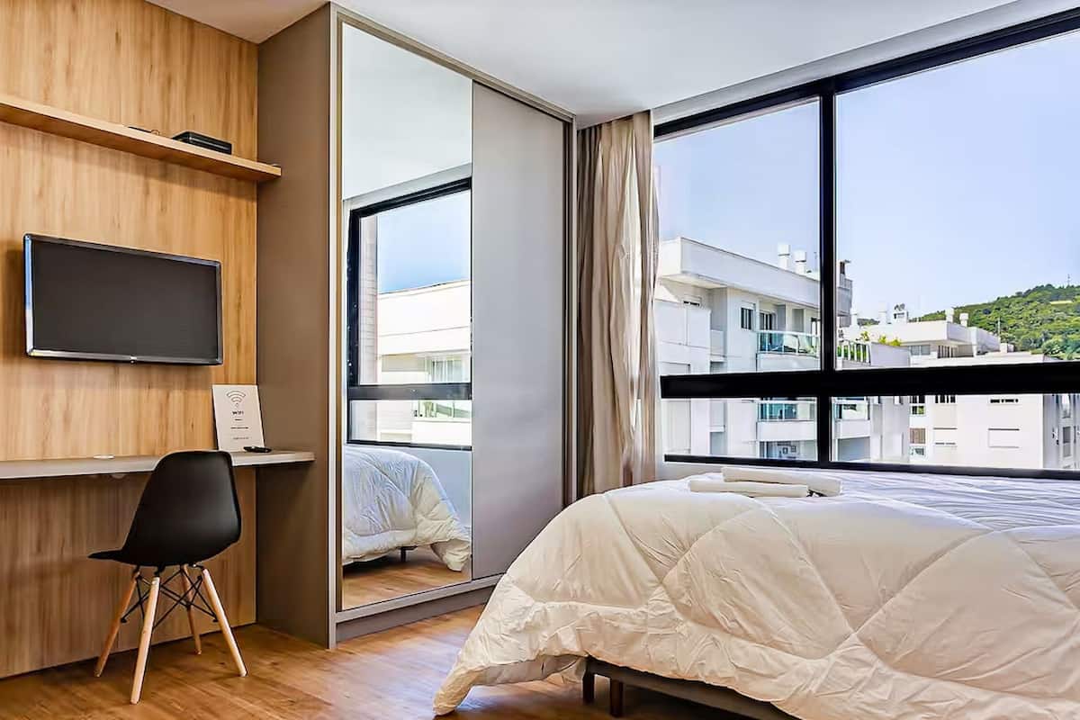 A modern hotel room with a large bed, a desk with a flat-screen TV, a chair, and a large window offering a city view.