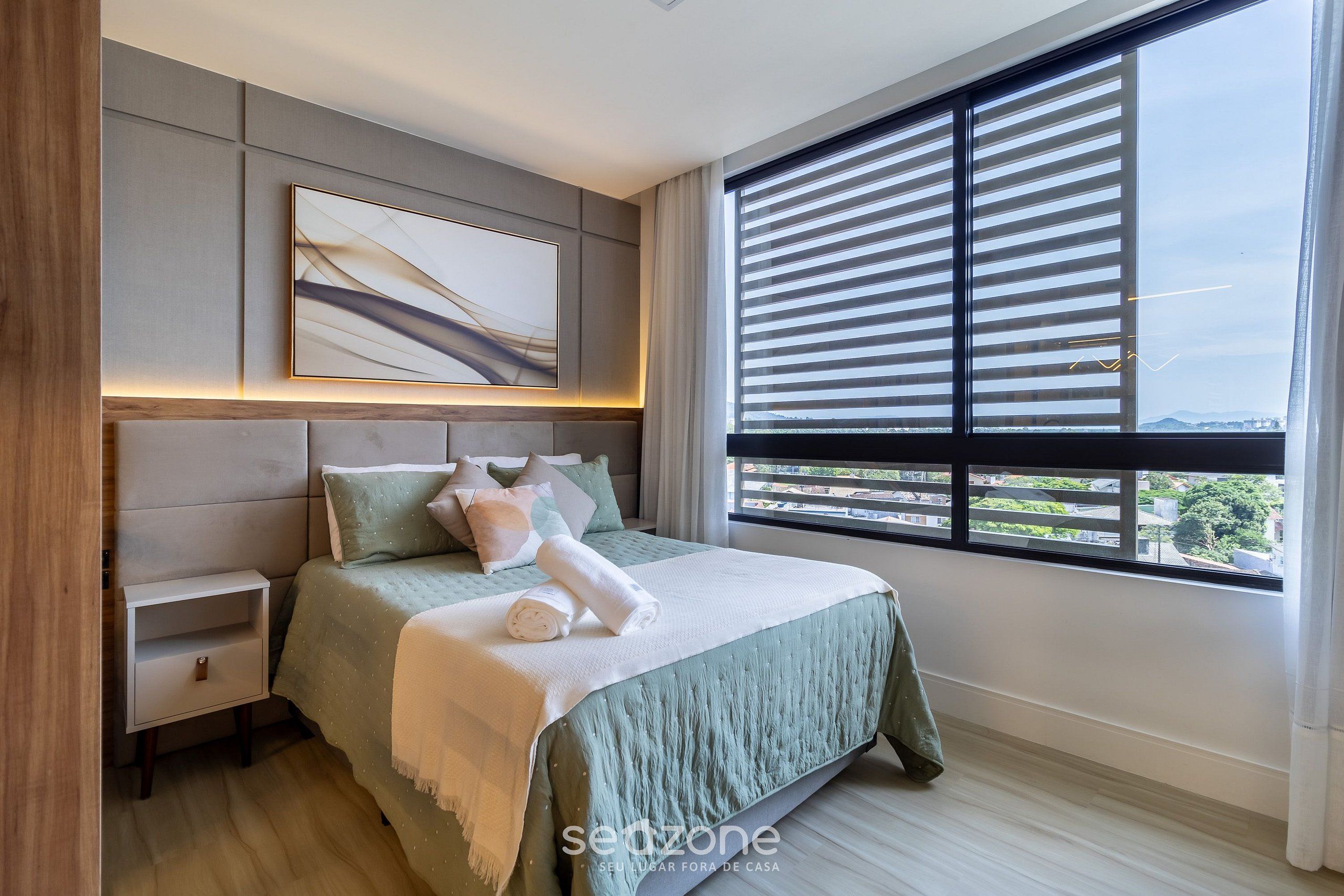 Un dormitorio moderno con una cama grande, una mesita de noche, una obra de arte en la pared y un ventanal con persianas.
