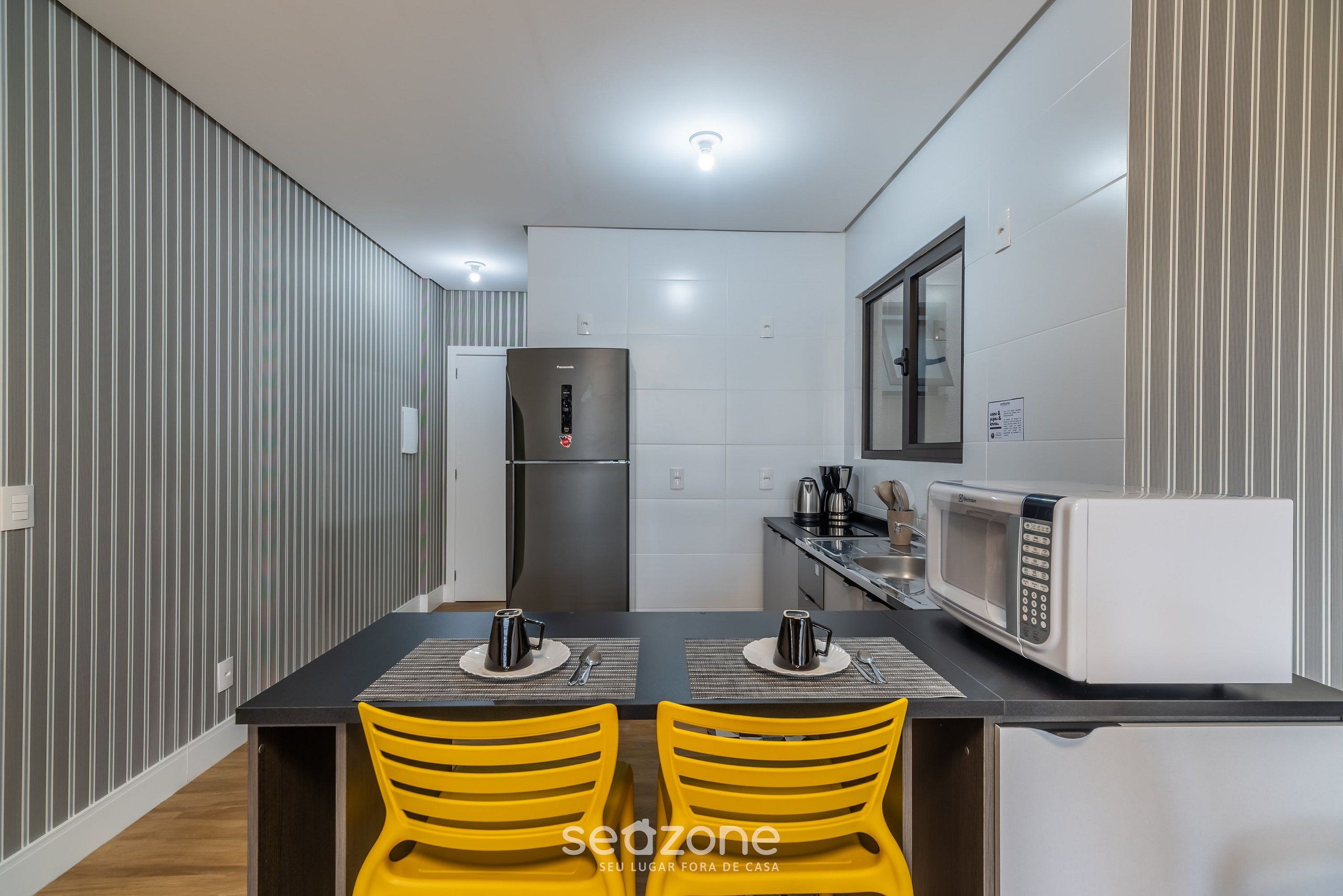 A modern kitchen with a refrigerator, microwave, and dining area with yellow chairs.