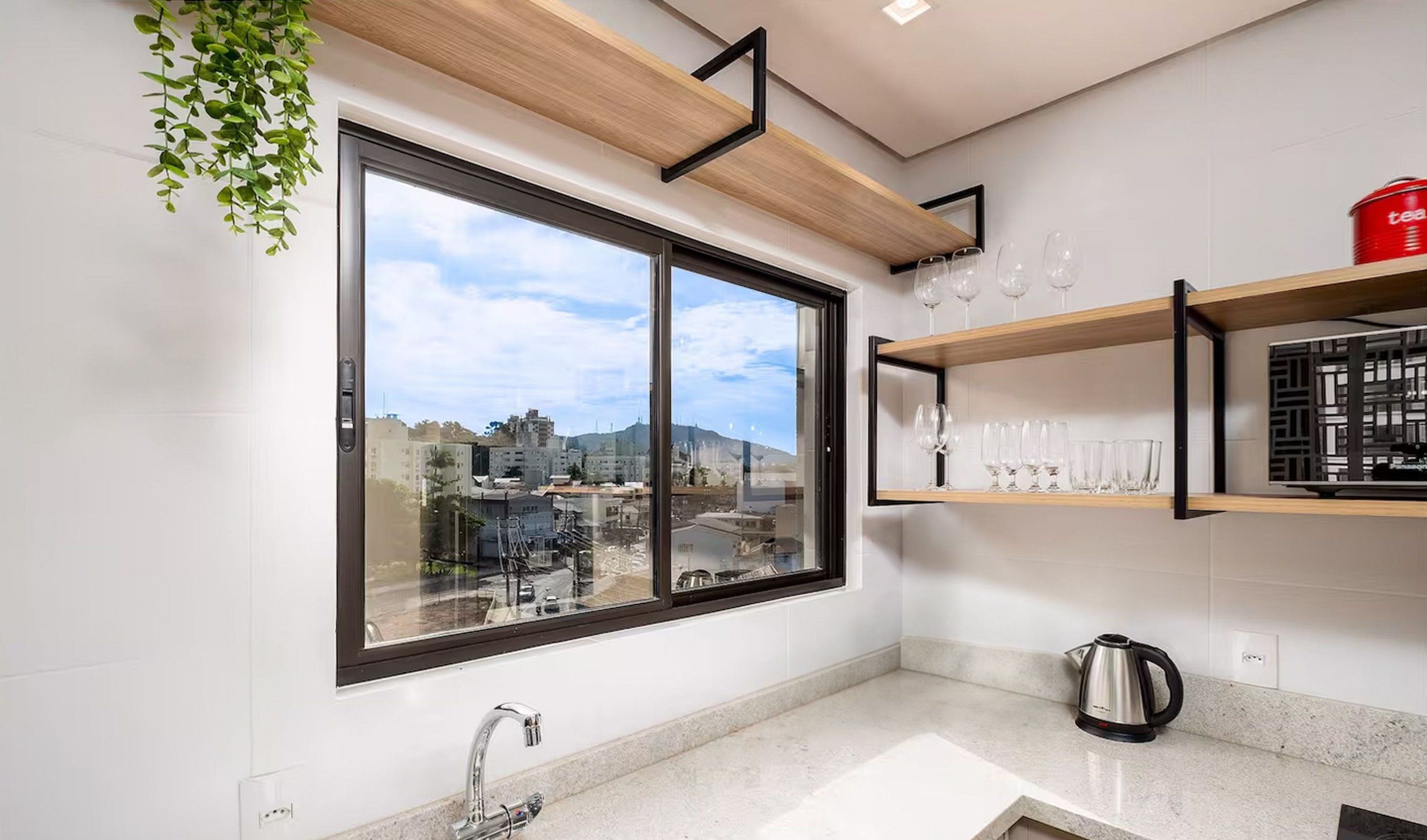 A modern kitchen with a large window, a stainless steel kettle, and wine glasses on shelves.