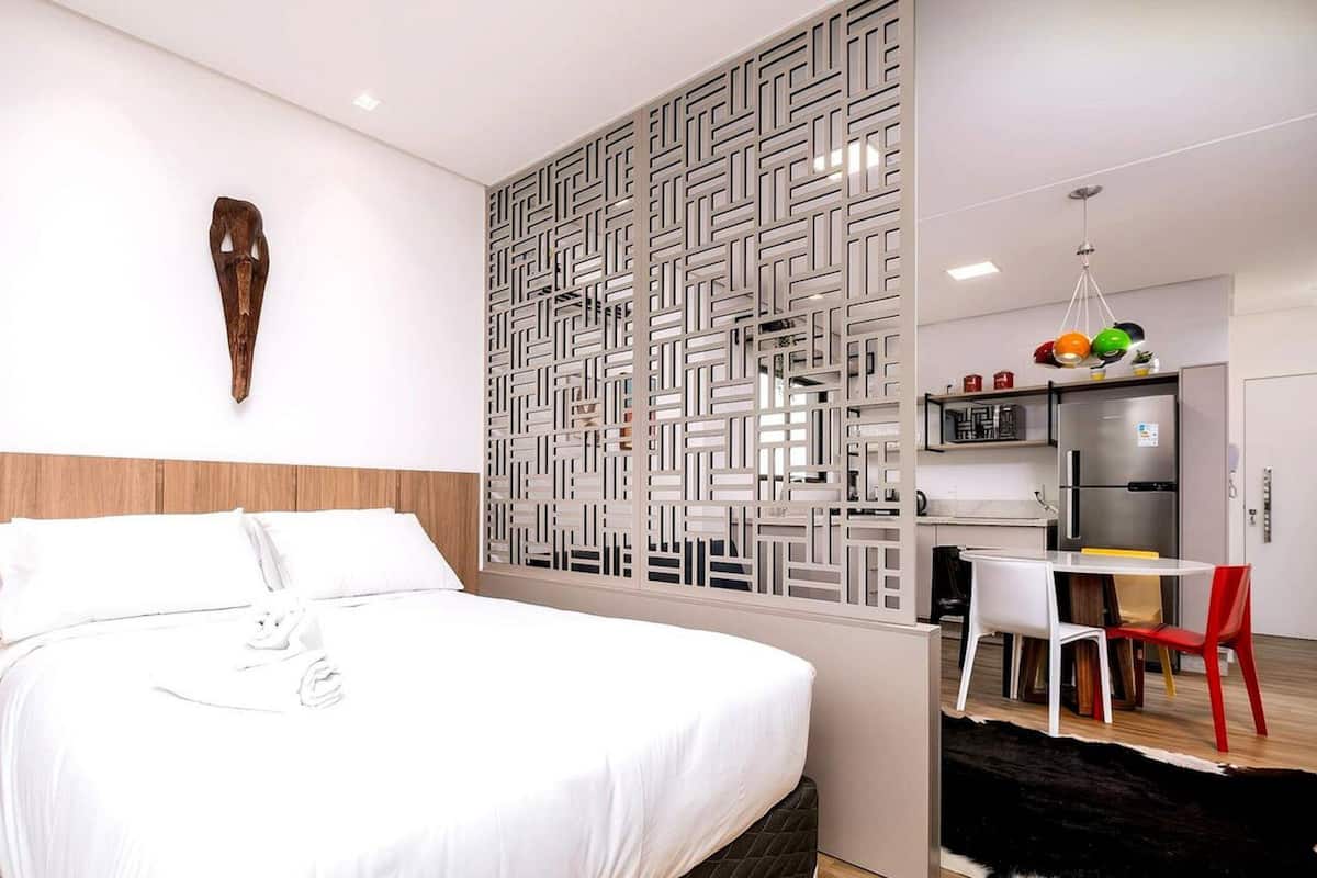 A modern hotel room with a large bed, a wooden headboard, a decorative wall panel, a small kitchen area with a refrigerator and a dining table with chairs, and a pendant light above the table.