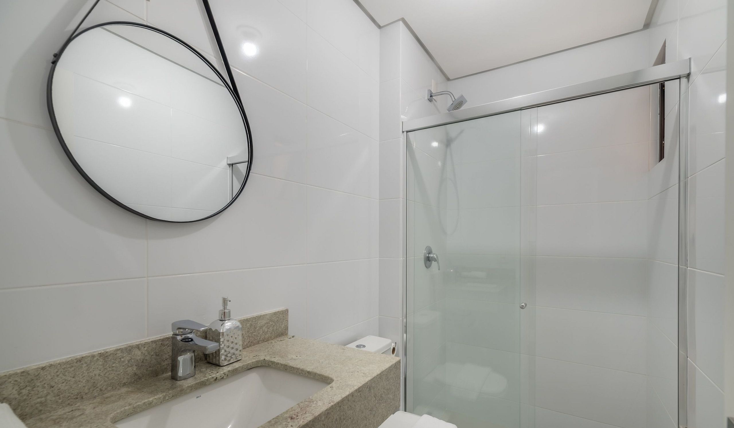 A modern bathroom with a glass shower enclosure, a round mirror, and a granite countertop with a sink.