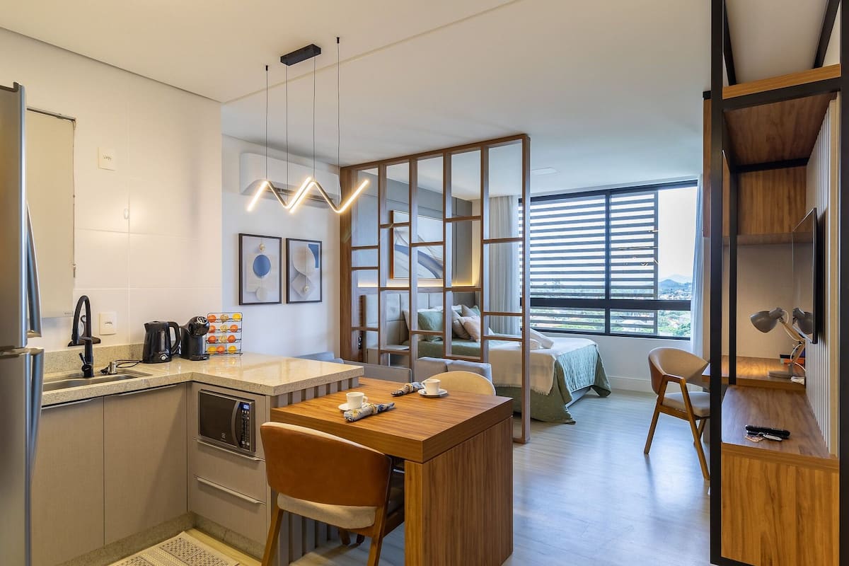A modern kitchen with a built-in oven, a coffee maker, and a kettle. There is a dining area with a wooden table and chairs.