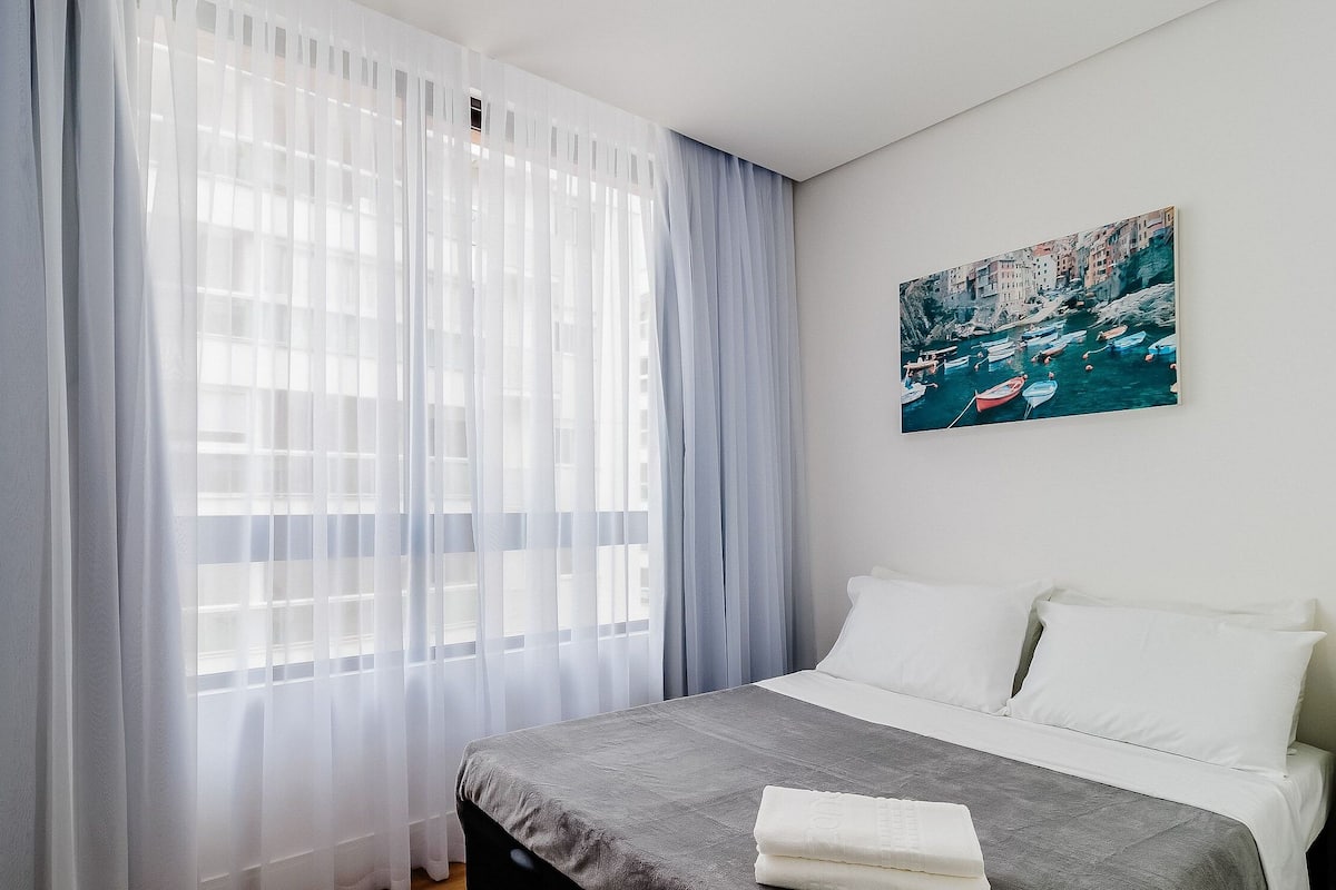 A hotel room with a bed, a painting of boats on the wall, and sheer curtains on the window.