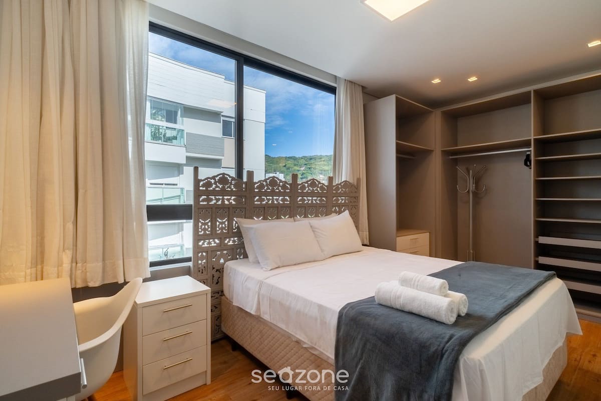 A bedroom with a large window, a bed with white bedding, a bedside table, and a wardrobe.