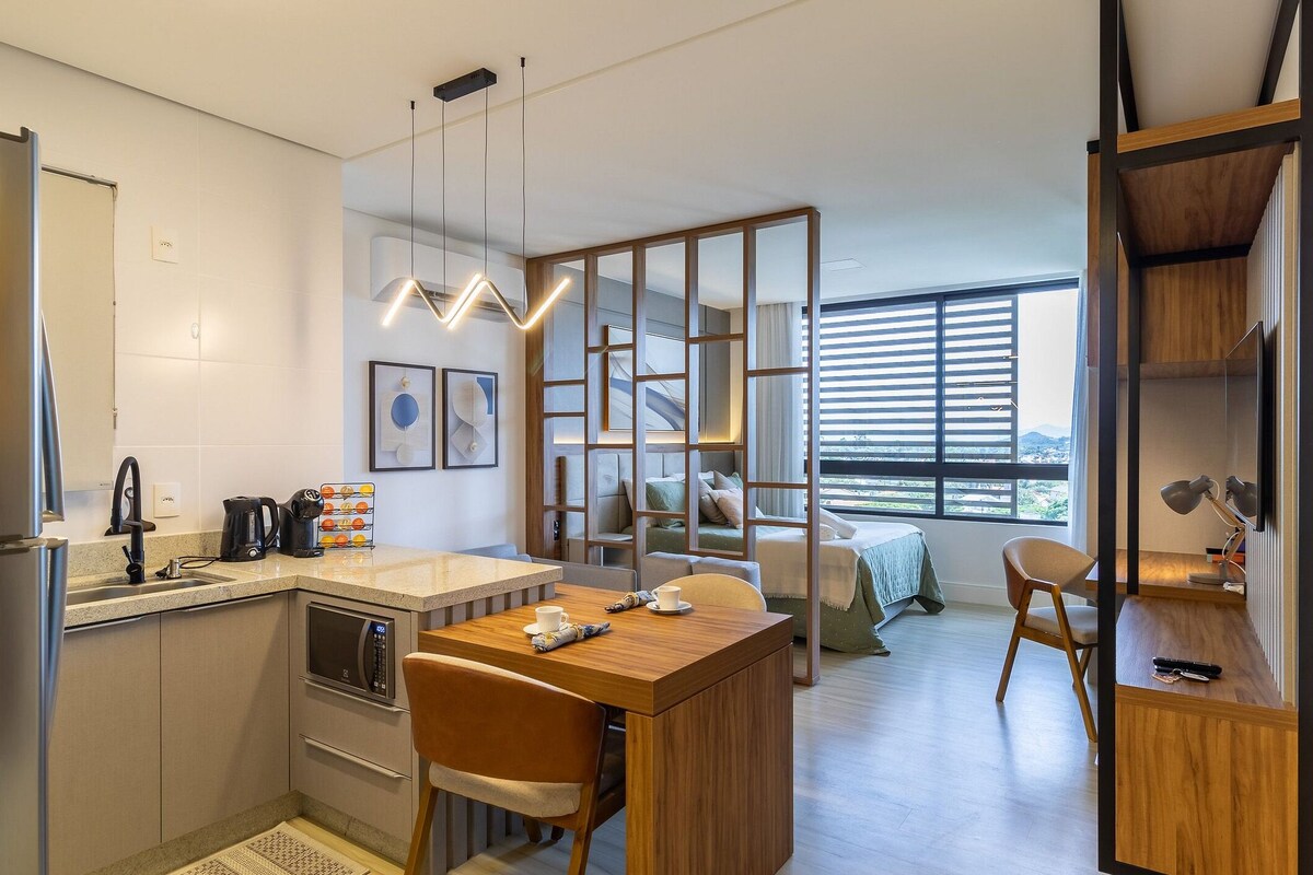 A modern kitchen with a built-in oven, a coffee maker, and a kettle. There is a dining area with a wooden table and chairs.