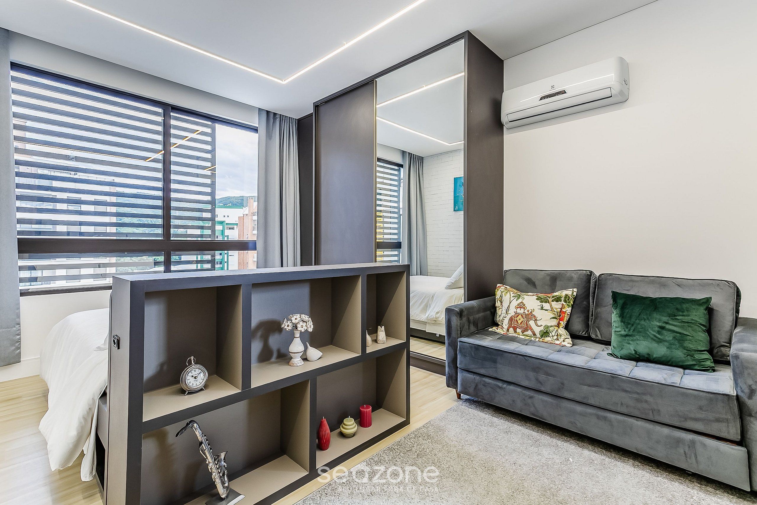 A modern living room with a grey sofa, a mirrored wardrobe, and a large window with blinds.
