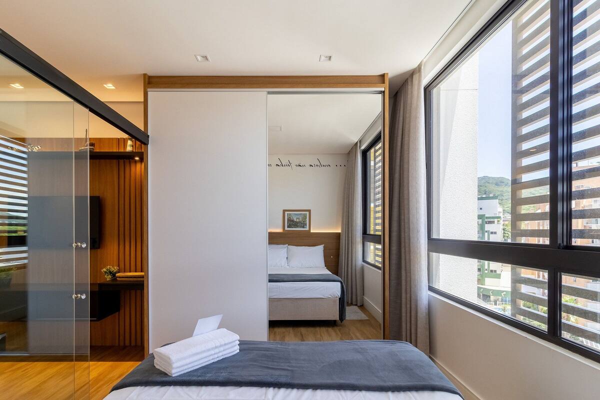 A modern hotel room with a large window, a bed, a bedside table, and a glass partition.
