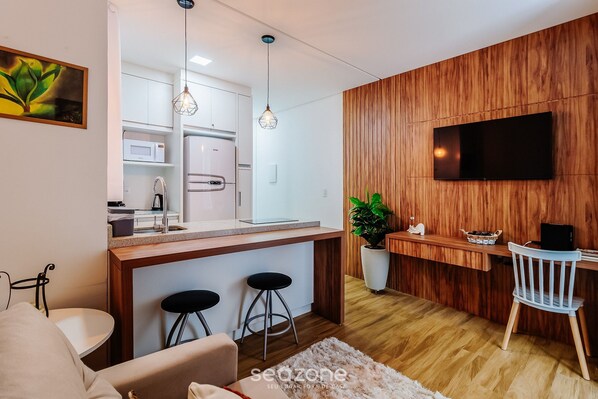 A modern kitchen with a wooden accent wall, a flat-screen TV, and a dining area with bar stools. - SSG Estúdios próximos a UFSC em Florianópolis (Florianópolis)