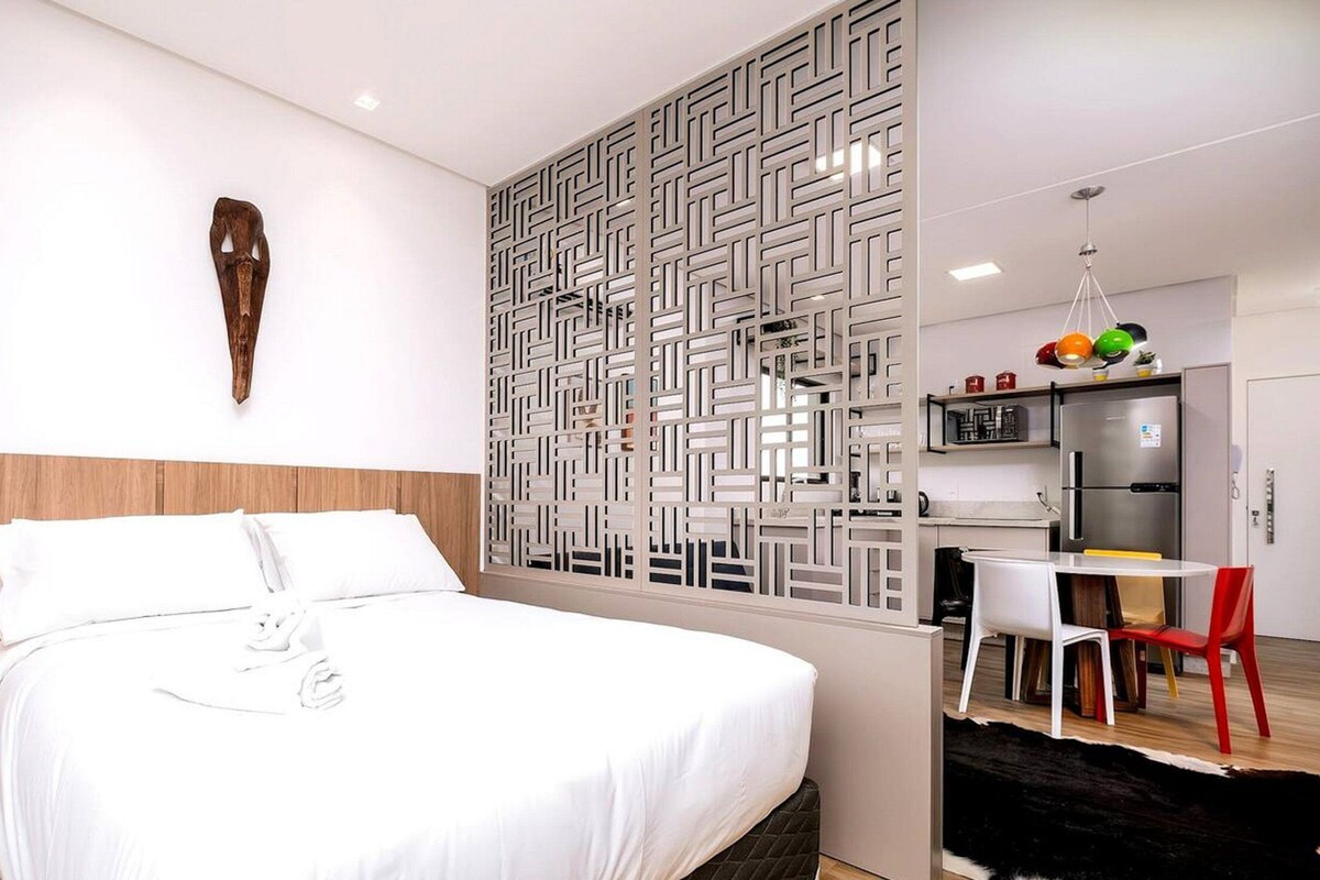 A modern hotel room with a large bed, a wooden headboard, a decorative wall panel, a small kitchen area with a refrigerator and a dining table with chairs, and a pendant light above the table.