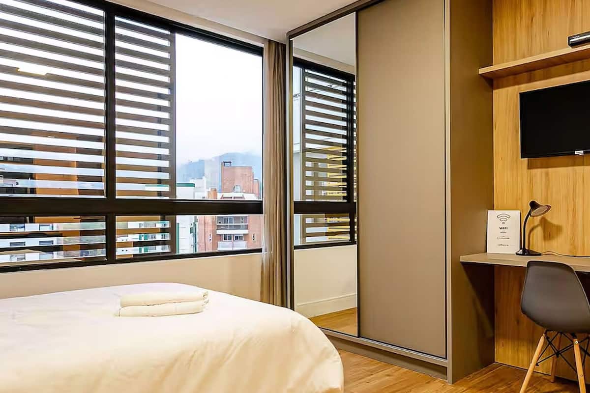 A hotel room with a bed, a desk, a chair, a TV, and a view of the city through large windows.