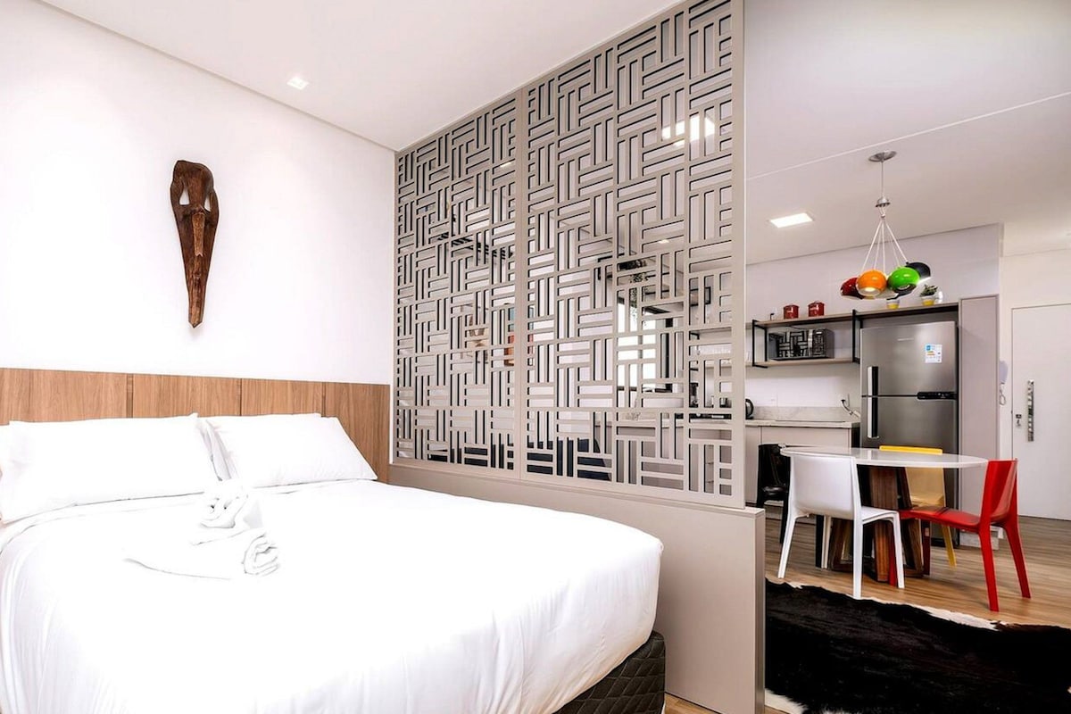 A hotel room with a bed, a desk, a chair, a kitchen area, and a decorative wall piece.