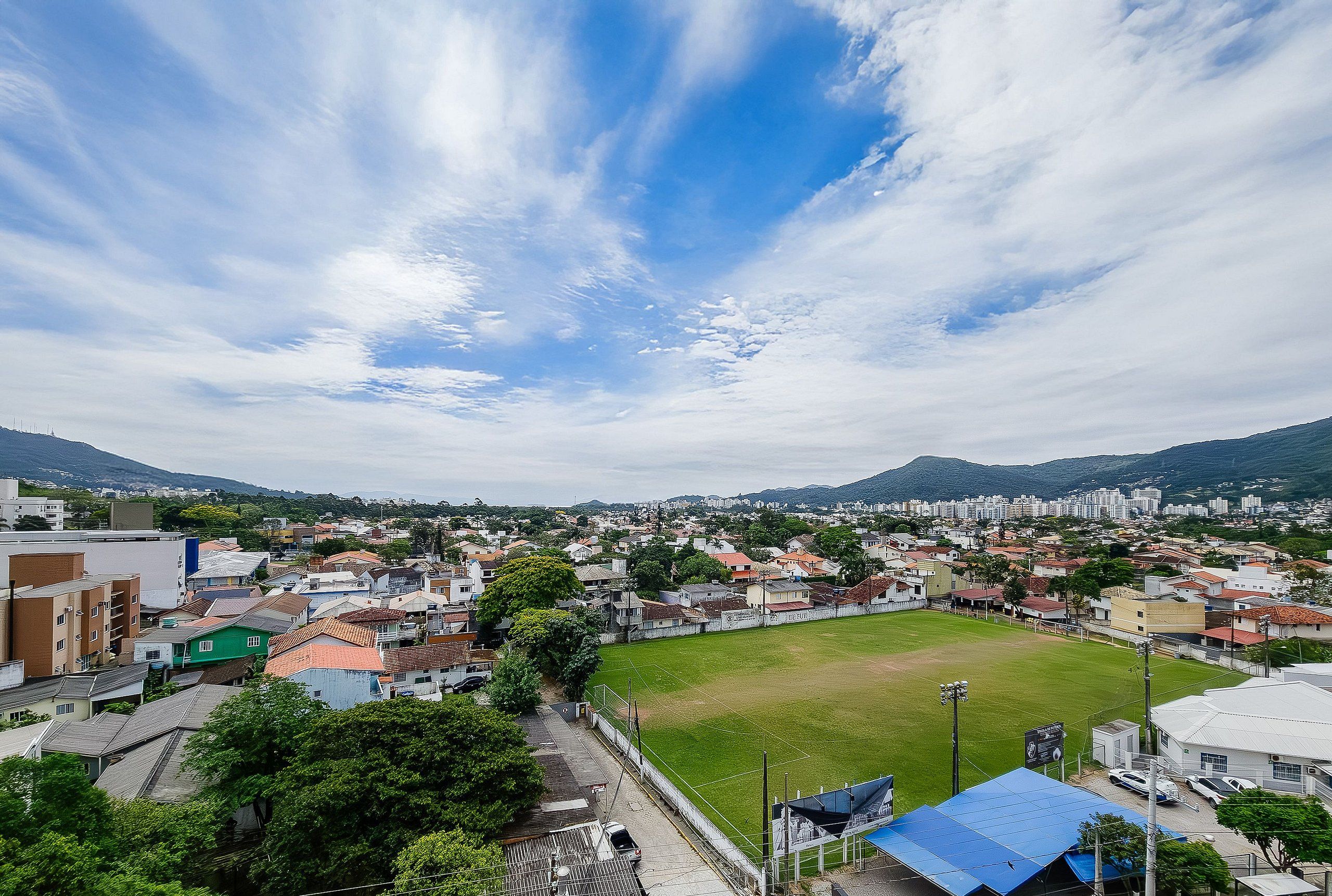 A panoramic view of a city with a large open field in the foreground, surrounded by residential buildings and a distant mountain range.
