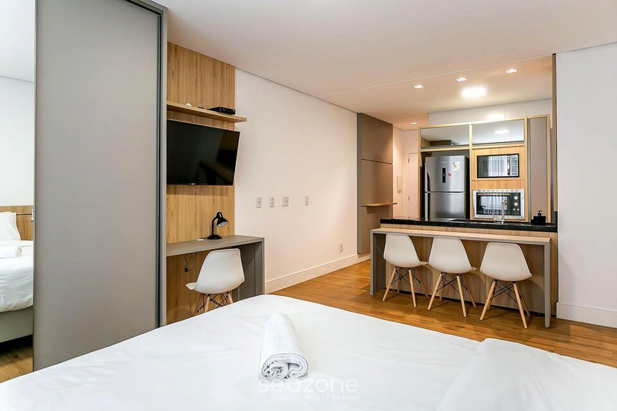 A modern hotel room with a bed, a desk, a TV, a small kitchenette, and a seating area with chairs.
