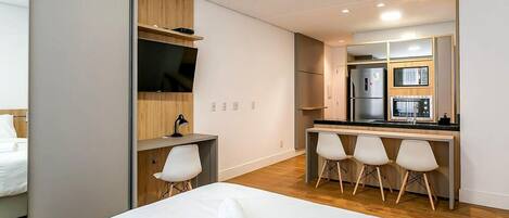 A modern hotel room with a bed, a desk, a TV, a small kitchenette, and a seating area with chairs.