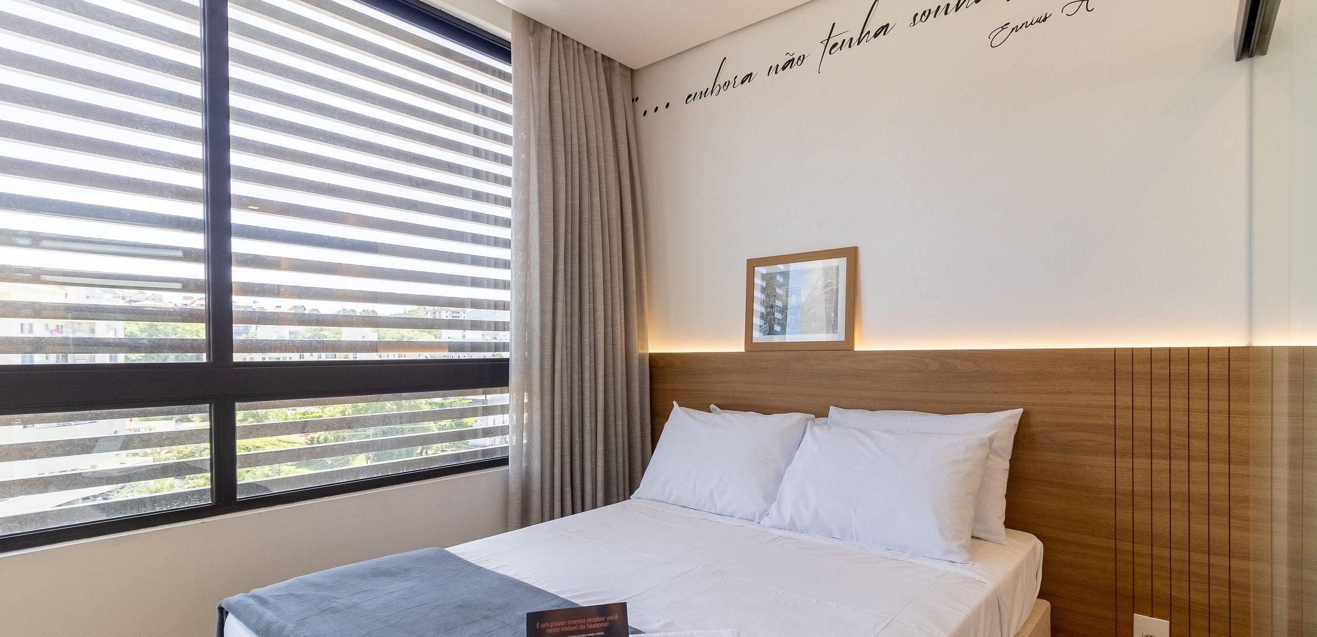 Una habitación de hotel con una cama, un ventanal con persianas, una cortina y una pared con un cuadro enmarcado.